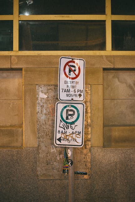 A vertical metal parking restriction sign mounted on a wall near a building with large glass windows at the top. The upper sign features a no-parking symbol with a red circle and slash over a black 'P