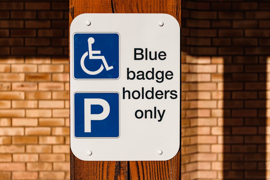 A vertical metal parking restriction sign mounted on a wall near a building with large glass windows at the top. The upper sign features a no-parking symbol with a red circle and slash over a black 'P